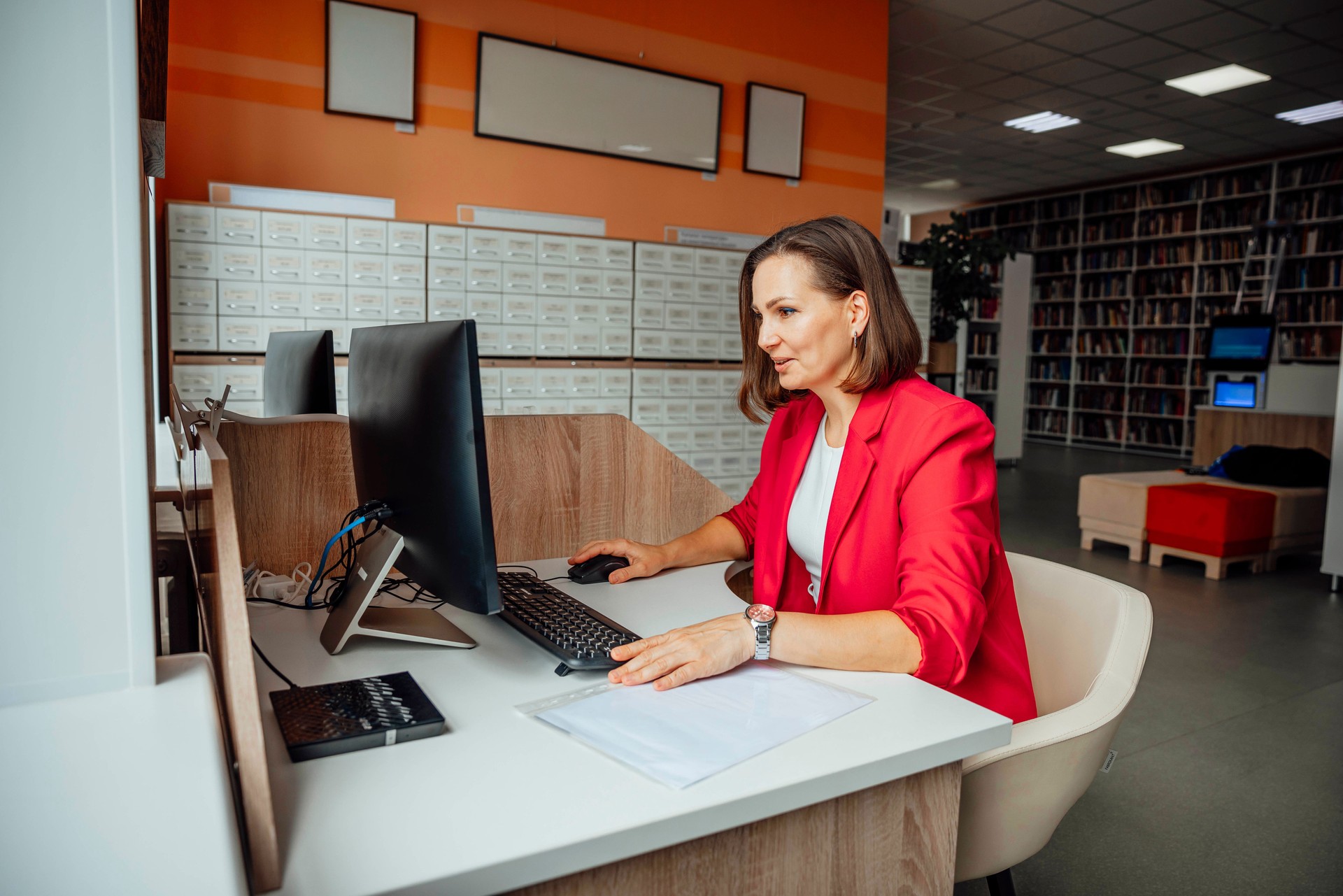 A young Caucasian woman with brown hair works at a computer in a modern coworking office. The space features wooden furniture and a library in the background.